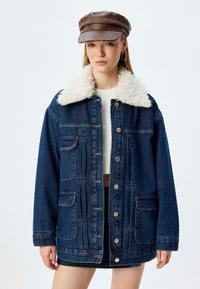 Dżinsowa kurtka z kołnierzem z owczej wełny, w ciemnoniebieskim kolorze, z frontowymi kieszeniami i zapięciem na guziki. Nosi się ją na jasnym swetrze i spódnicy.