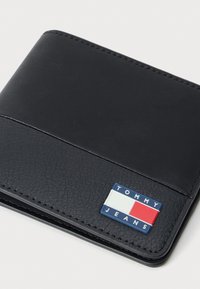 Μαύρο δερμάτινο πορτοφόλι με υφή, που διαθέτει μια μπλε και κόκκινη επωνυμία με την ένδειξη "TOMMY JEANS" στο μπροστινό μέρος.