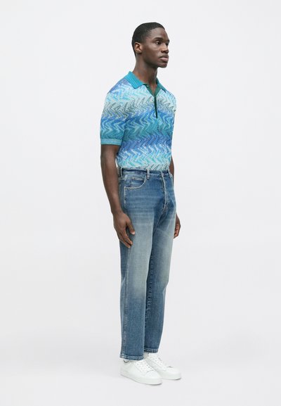 Kék rövid ujjú galléros póló hullámmintával, világoskék, egyenes szárú, kifakult farmernadrággal és fehér sportcipővel párosítva.