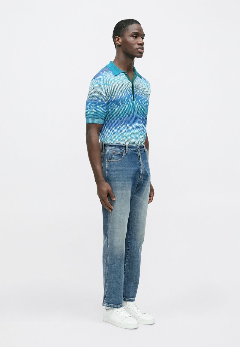 Polo à manches courtes bleu avec motif de vagues, associé à un jean bleu délavé à jambes droites et des baskets blanches.