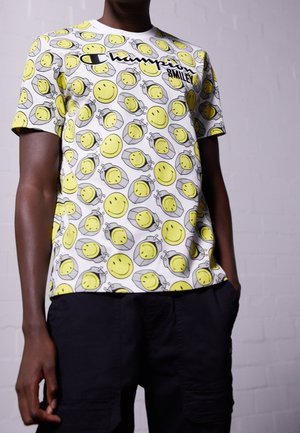 Homme portant un t-shirt blanc Champion couvert de visages souriants jaunes et un short noir avec les mains dans les poches.
