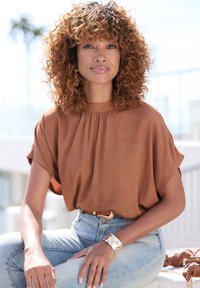 Braunes kurzärmliges Oberteil mit gerafftem Ausschnitt, kombiniert mit hoch taillierten hellblauen Jeans. Ein goldenes Armreif und ein Gürtel akzentuieren das Outfit.