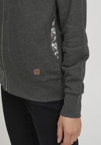 Felpa grigia con zip, dettagli sul taschino a fantasia floreale, texture morbida e polsini a costine. Piccolo etichetta logo marrone vicino all'orlo.