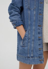 Veste en denim bleu à coupe décontractée, avec manches retroussées, poches avant et boutons métalliques sur la fermeture. Couche en maille confortable en dessous.