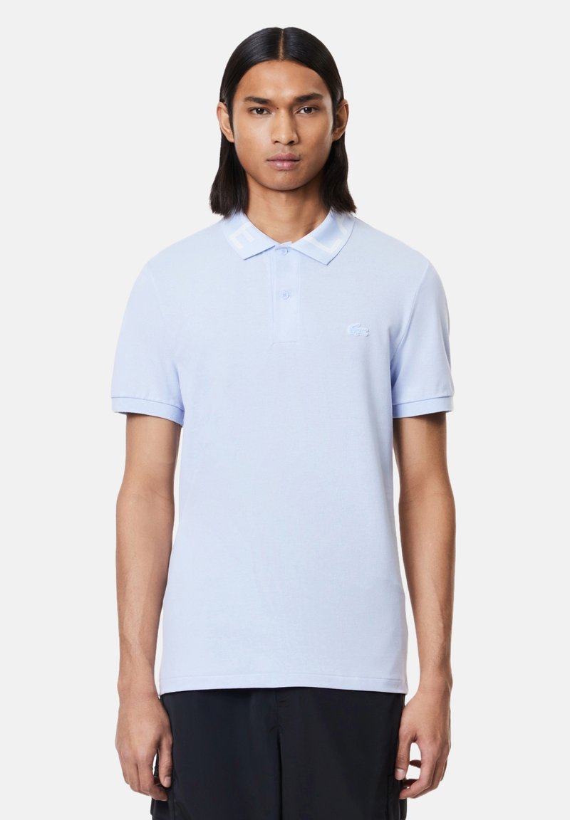 Camisa polo azul claro con collar clásico, mangas cortas y un pequeño logo bordado en el pecho. Textura de tejido suave y lisa.
