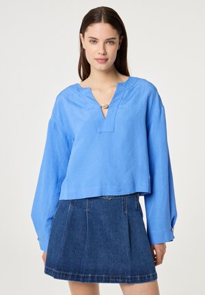 Vrouw die een felblauwe blouse met lange mouwen draagt, met een V-hals met knopen, en een donkerblauwe denim minirok, tegen een effen achtergrond.