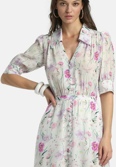 Femme portant une robe blanche à fleurs roses et violettes, manches bouffantes, boutonnée à l'avant, boucles d'oreilles anneaux et bracelets blancs au poignet gauche.