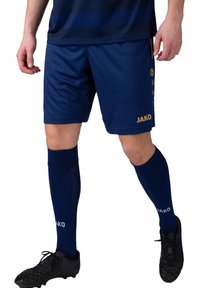 Shorts de sport navy associés à des chaussettes navy jusqu'aux genoux. Présente un logo doré "JAKO" et un motif texturé sur des chaussures de football noires.