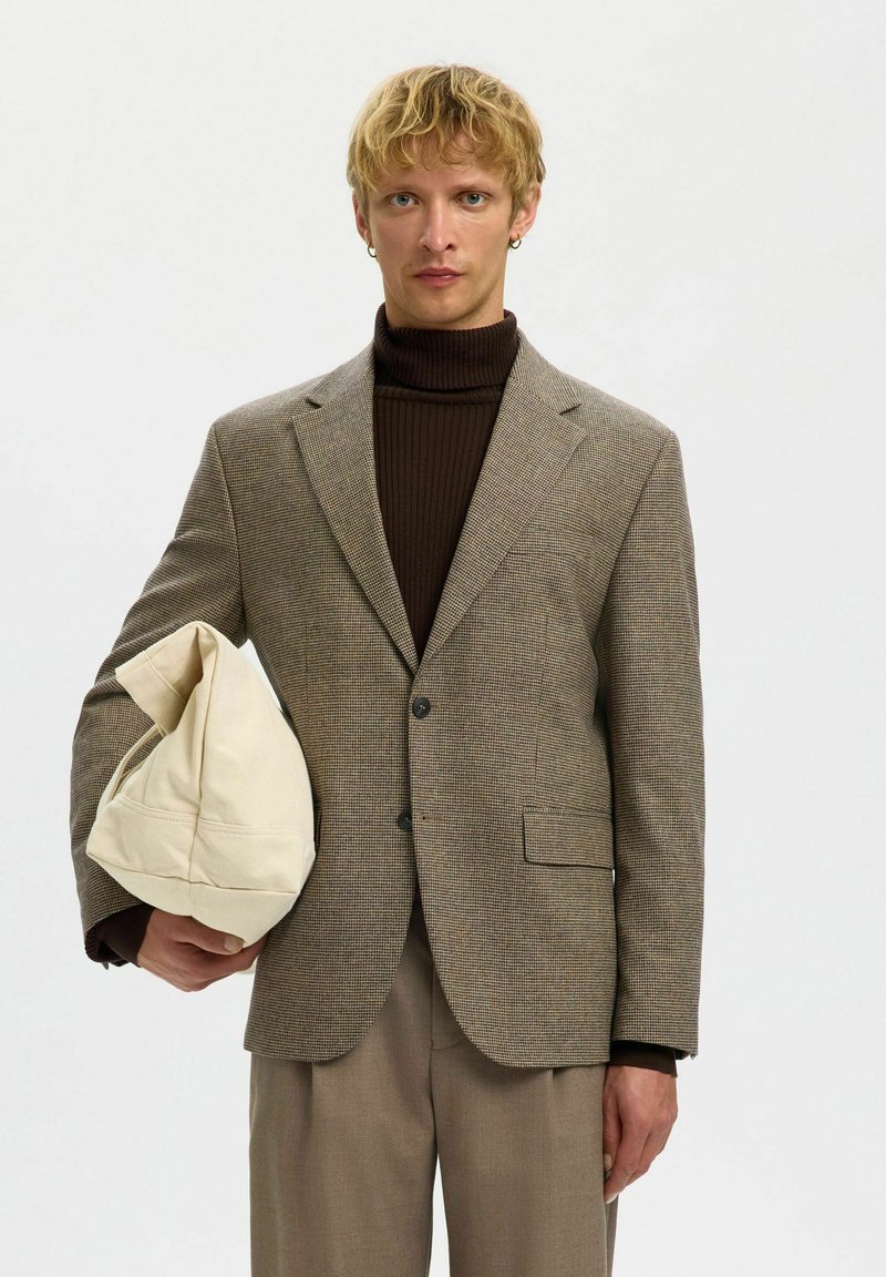 Brun rutete blazer med skreddersydd passform, enkelt knappelukking, båret over en mørk høyhalset genser. Modellen holder en kremfarget veske.