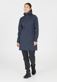 Mörkblå lång kappa med dragkedja framtill och huva, tillverkad av slät tyg. Har sidofickor och en rak passform, tillsammans med svarta byxor och stövlar.