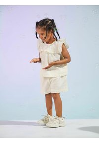 Hvid, kortærmet bluse med flæser og hulmønster, parret med matchende shorts. Modellen bærer lyse sneakers og er omgivet af sæbebobler.