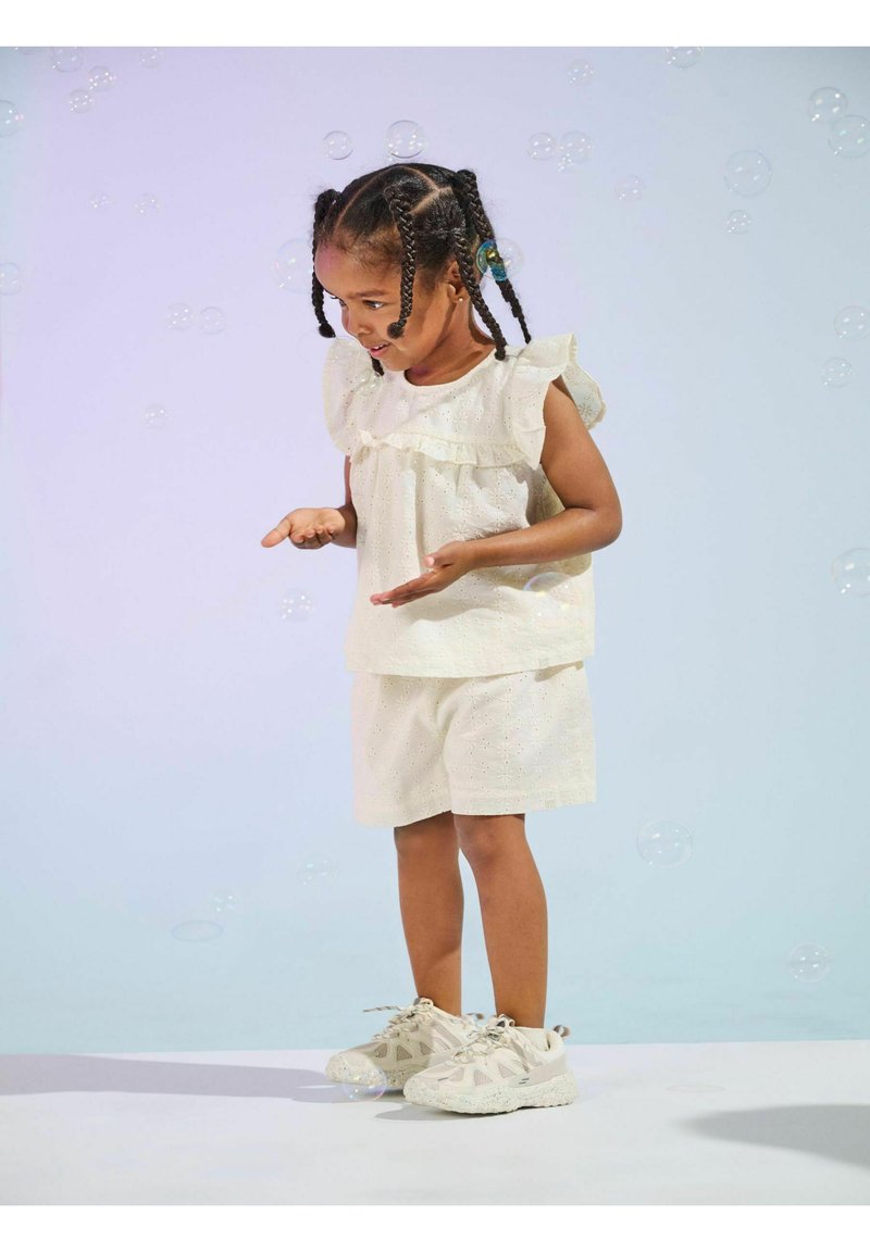 Hvid, kortærmet bluse med flæser og hulmønster, parret med matchende shorts. Modellen bærer lyse sneakers og er omgivet af sæbebobler.