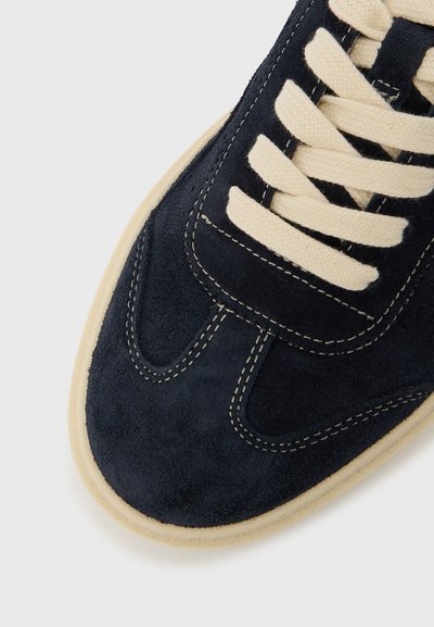 Tengerészkék velúr sportcipő kerek orral, krémszínű fűzőkkel és kontrasztos bézs gumitalppal. Látható varrással és sima textúrával rendelkezik.