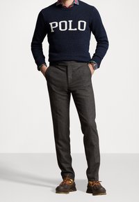 Marinblå stickad tröja med vit "POLO"-logotyp, lager på en rutig skjorta. Grå skräddarsydd byxor och bruna skor med snören fulländar outfiten.