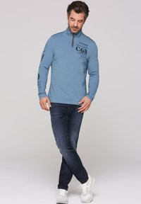 Maglia a maniche lunghe zip-up di colore blu chiaro con testo stampato, abbinata a jeans scuri e sneaker bianche. La maglia presenta un colletto alto e cuciture a contrasto.