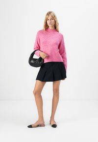 Mulher a usar um suéter rosa, uma saia mini plissada preta, uns sapatilhas de tiras bicolores, a segurar uma pequena mala preta contra um fundo branco.