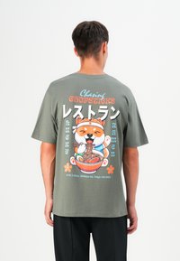 Olijfgroen T-shirt met korte mouwen, voorzien van een kleurrafische afbeelding van een hond met chopsticks en ramen, aangevuld met tekst en bloemmotieven.