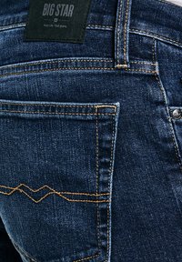 Tumesinised teksapüksid tagataskuga, oranži õmblusega ja musta "BIG STAR" kaubamärgiga silt vööjoone kohal.