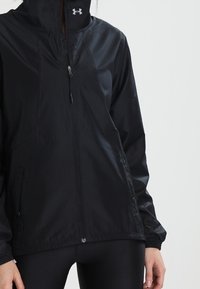 Chaqueta negra ligera con un cuello alto, cierre frontal de cremallera y puños elásticos. Presenta un sutil logotipo en el pecho y una textura suave.