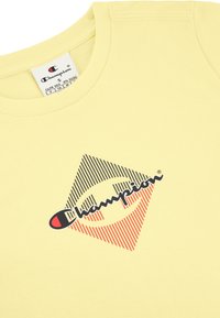 Camiseta de algodón amarilla con cuello redondo, que presenta un logo de diamante a rayas rojas y azules y texto negro en la parte frontal.