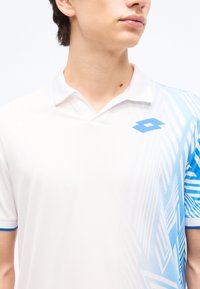 Νεαρός που φοράει άσπρο πόλο μπλουζάκι με μπλε γεωμετρικό σχέδιο στον δεξί ώμο και μπλε λογότυπο στο στήθος.