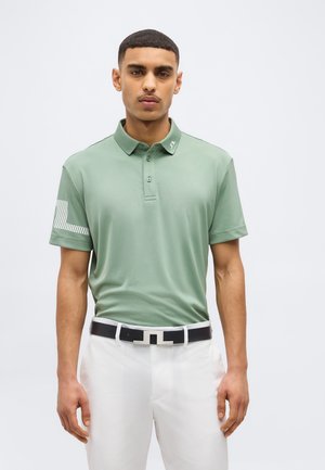 Mann iført lys grønn polotrøye med tre knapper og hvite bukser med svart-hvitt belte, stående mot en ensfarget bakgrunn.