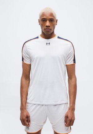 Άνδρας με κοντά ξανθισμένα μαλλιά που φοράει λευκό αθλητικό πουκάμισο και σορτς με λεπτομέρειες σε ναυτικό μπλε και πορτοκαλί, στέκεται μπροστά σε απλό φόντο.