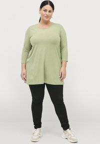 Tunique verte claire à manches trois-quarts, coupe ample, associée à un jean skinny noir et des baskets blanches avec des accents beiges.