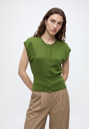 Femme aux longs cheveux bruns portant un haut vert sans manches boutonné et un pantalon beige taille haute sur un fond clair uni.
