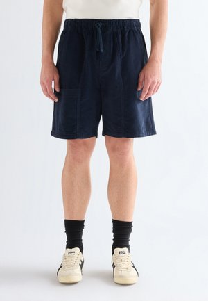 Persona che indossa pantaloncini con coulisse blu navy, calzini neri alla caviglia e scarpe da ginnastica bianche, in piedi contro uno sfondo semplice.