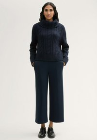 Marineblauer Kabelstrickpullover mit weitem Rollkragen, kombiniert mit dunkelgrauen, weiten Hosen und schwarzen Schnürschuhen. Glatte Textur und bequeme Passform.