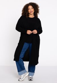 Schwarzer langer Cardigan mit offener Vorderseite, zwei Taschen, gerippten Bündchen und einer weichen Textur, getragen über einem schwarzen Oberteil und kombiniert mit blauen Jeans und silbernen Sneakern.