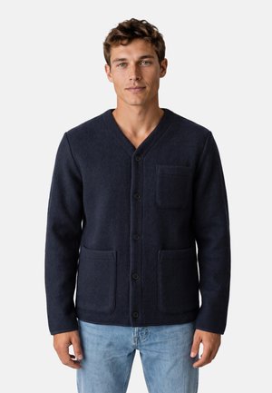 Giovane uomo che indossa un cardigan blu navy scuro abbottonato con tre tasche e jeans azzurri, in piedi davanti a uno sfondo semplice.