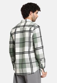 Camicia a quadri verde e bianca con dettagli neri, maniche lunghe, colletto a bottoni e orlo curvo. Tessuto morbido e testurizzato.