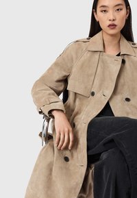 Μπεζ δερμάτινο παλτό τρανς με διπλό πέτο, μεγάλους γιακάδες, μπροστινές τσέπες και λεπτομέρειες με κουμπιά, φορεμένο πάνω από σκούρο υφασμάτινο παντελόνι.