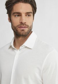 Witte poloshirt met een klassieke kraag, de stof heeft een subtiele textuur en een knoopsluiting aan de voorkant. Het model heeft lichtbruin haar en een baard.
