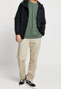 Person wearing a black hooded jacket, olive green t-shirt, beige pants, and black-and-white sneakers standing with one hand in pocket.