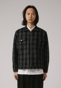 Veste en flanelle à carreaux noirs et gris avec poches avant, fermeture éclair et col, portée sur une chemise blanche et un pantalon noir.