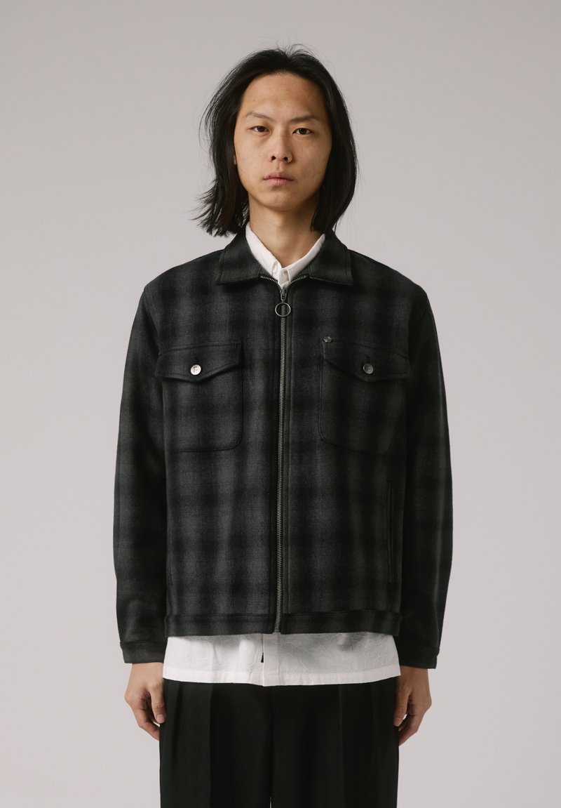 Veste en flanelle à carreaux noirs et gris avec poches avant, fermeture éclair et col, portée sur une chemise blanche et un pantalon noir.