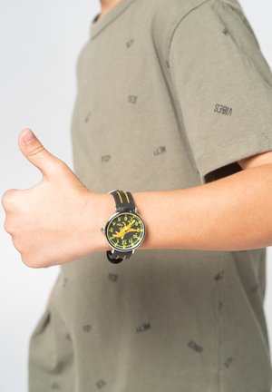 Montre analogique avec un boîtier rond noir, un cadran jaune et des inscriptions noires et jaunes en gras. Le bracelet est noir avec des rayures orange.