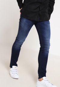 Jeans ajustados de un azul oscuro hechos de mezclilla, con una textura desgastada y detalles de costura mínimos. Combinados con zapatillas deportivas blancas.