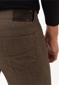 Brown denim jeans with a textured finish and a leather patch on the back waistband. Features a single back pocket with stitching detail.