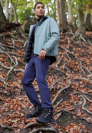 Veste d'extérieur bleu clair, associée à un pantalon marine et des bottes de randonnée noires. Présente un col haut, une fermeture éclair et un design ajusté.