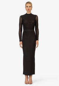 Robe noire à manches longues avec un haut transparent et texturé et une jupe unie. Présente une silhouette ajustée et une fente latérale ; portée avec des escarpins nude.