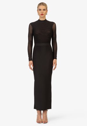 Robe noire à manches longues avec un haut transparent et texturé et une jupe unie. Présente une silhouette ajustée et une fente latérale ; portée avec des escarpins nude.