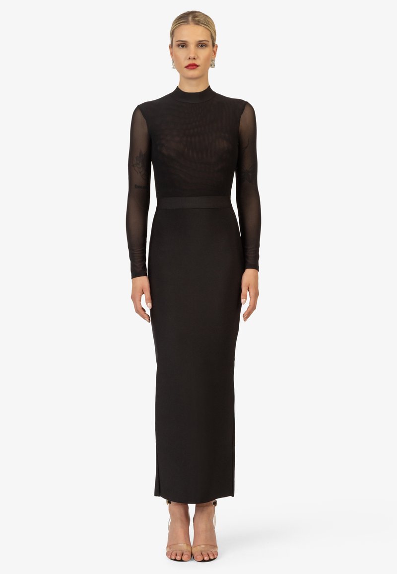 Robe noire à manches longues avec un haut transparent et texturé et une jupe unie. Présente une silhouette ajustée et une fente latérale ; portée avec des escarpins nude.