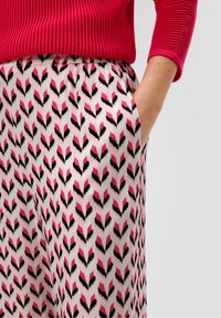 Mano nella tasca di una gonna a fantasia rosa, nera e bianca abbinata a una maglietta rossa a maniche lunghe a costine, su uno sfondo neutro.