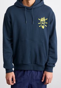 Tummansininen huppari, jossa on etutasku, säädettävä naruhuppu ja keltainen graafinen kuva ranskalaisista perunoista kupissa säteilevin viivojen kanssa.