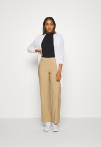 White knitted cardigan with lace details, black ribbed turtleneck, beige high-waisted trousers, and white sneakers, model stands with arms crossed.