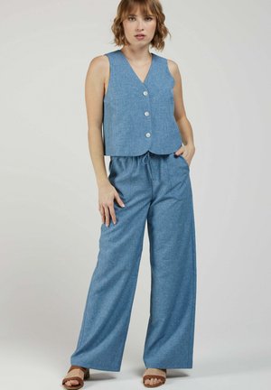 Femme aux cheveux courts portant un gilet bleu sans manches boutonné et un pantalon large assorti, debout avec une main dans la poche et des sandales marron.